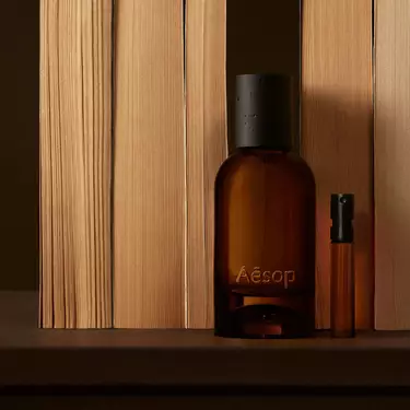 Amber Aesop bottle and slim vial placed between vertical pale wood boards, with deep brown shadows and a clean linear composition.