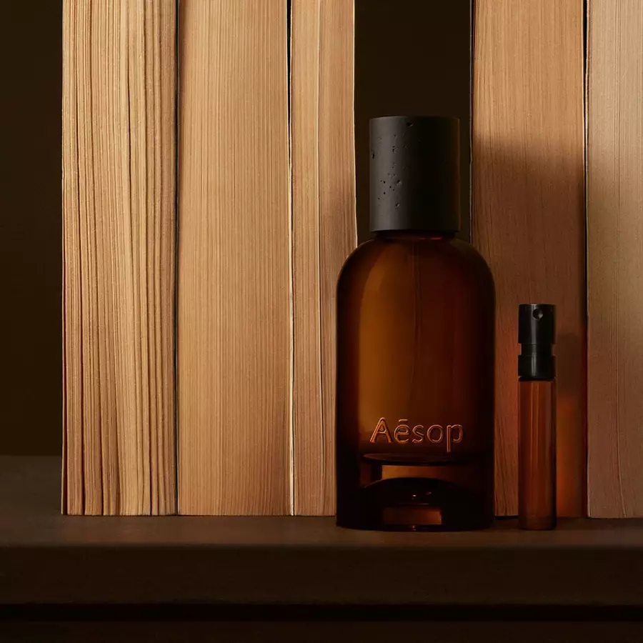 Aesop fragrance bottle and travel-size bottle in amber glass with black caps on a warm wooden shelf in soft light.