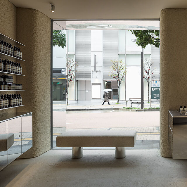 Industrial Aesop store design with stainless steel counters, minimalist bench, and a city street view