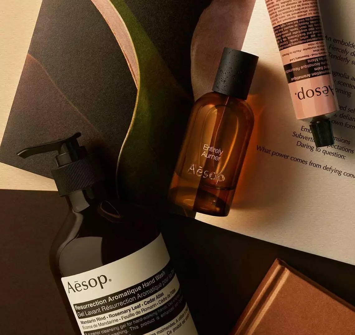 Aesop Entirely, Aurner fragrance bottle, large pump bottle and cream tube on a surface with printed material.