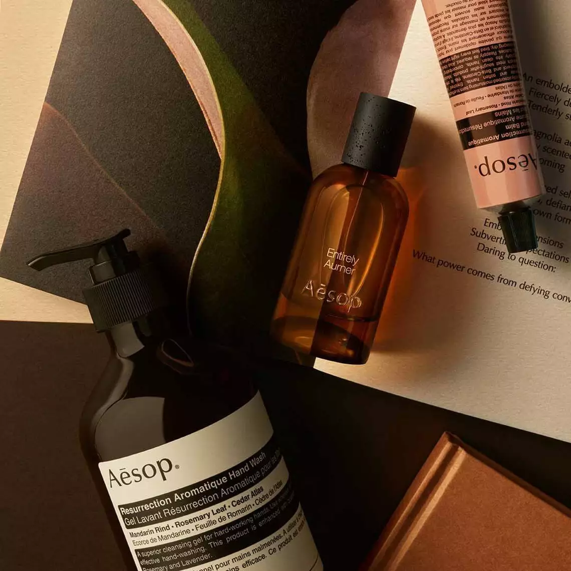 Aesop Entirely, Aurner fragrance bottle, large pump bottle and cream tube on a surface with printed material.