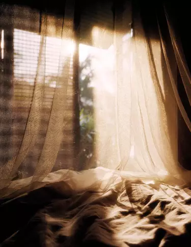 Sheer draped curtains over rumpled fabric near a sunlit window, showing warm beige tones, soft shadows, and translucent folds.