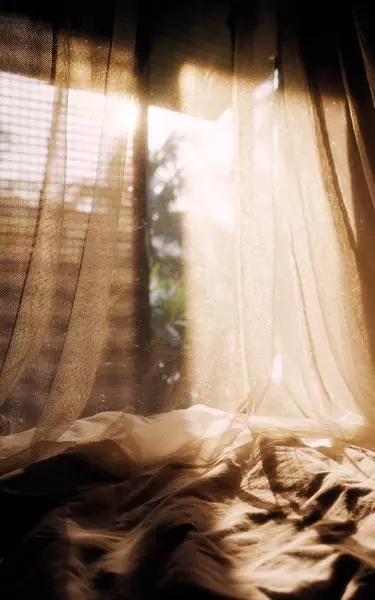 Sheer draped curtains over rumpled fabric near a sunlit window, showing warm beige tones, soft shadows, and translucent folds.