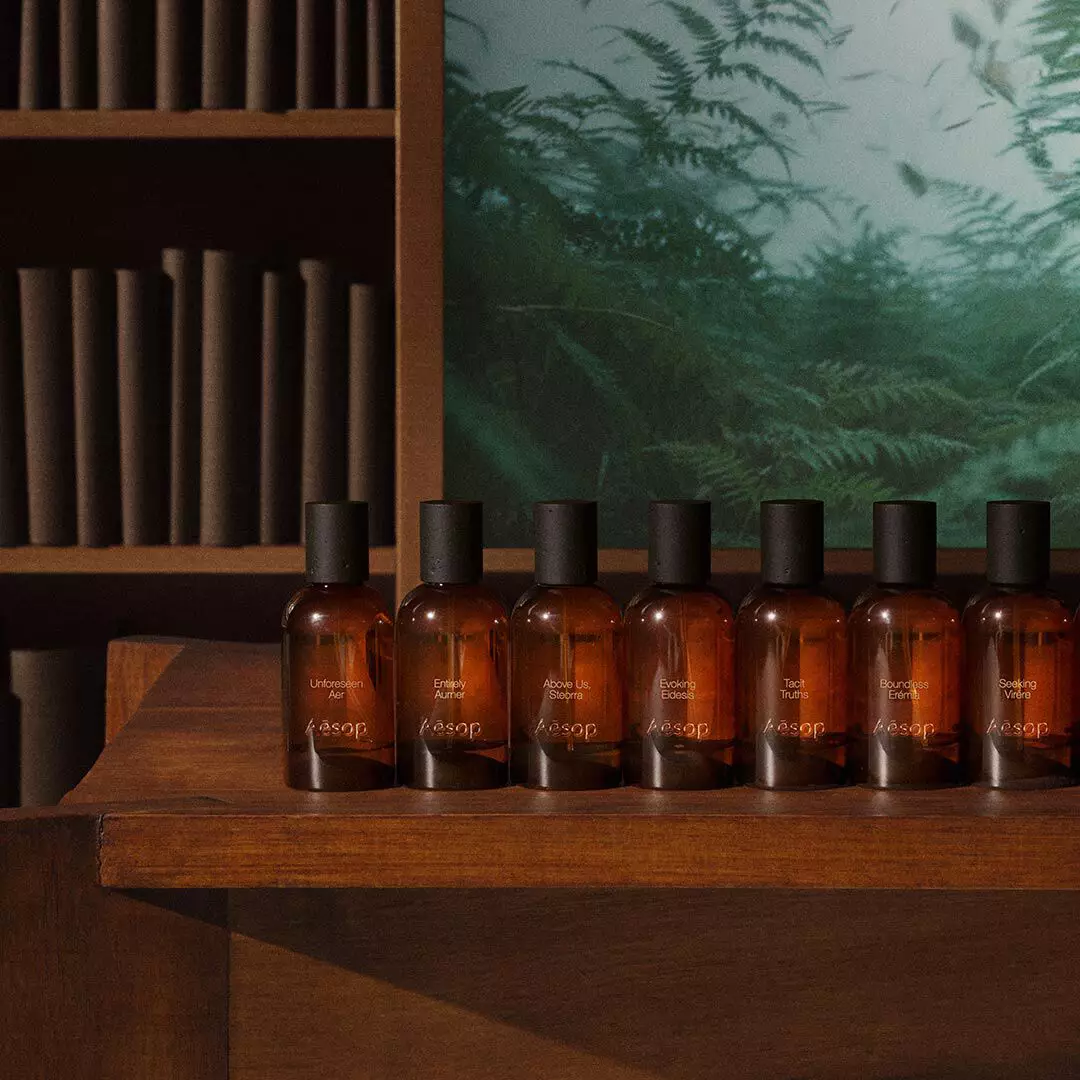Row of amber Aesop fragrance bottles on a wooden table beside shelves and a green fern image.
