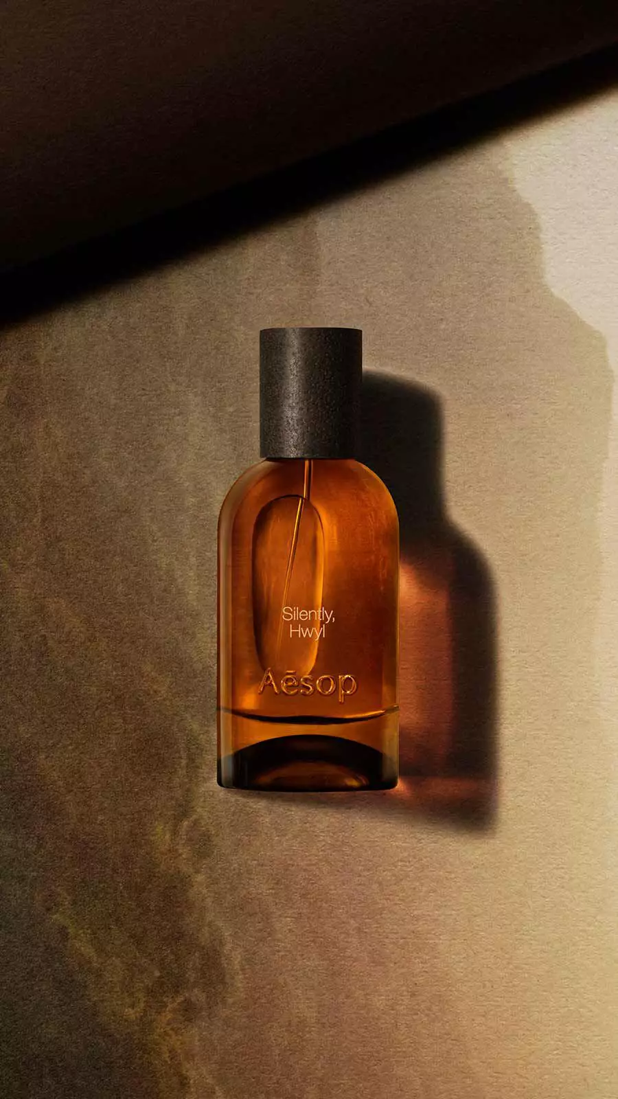 Aesop Silently, Hwyl Eau de Parfum bottle with black cap against a warm golden-brown stone-textured wall with soft light.