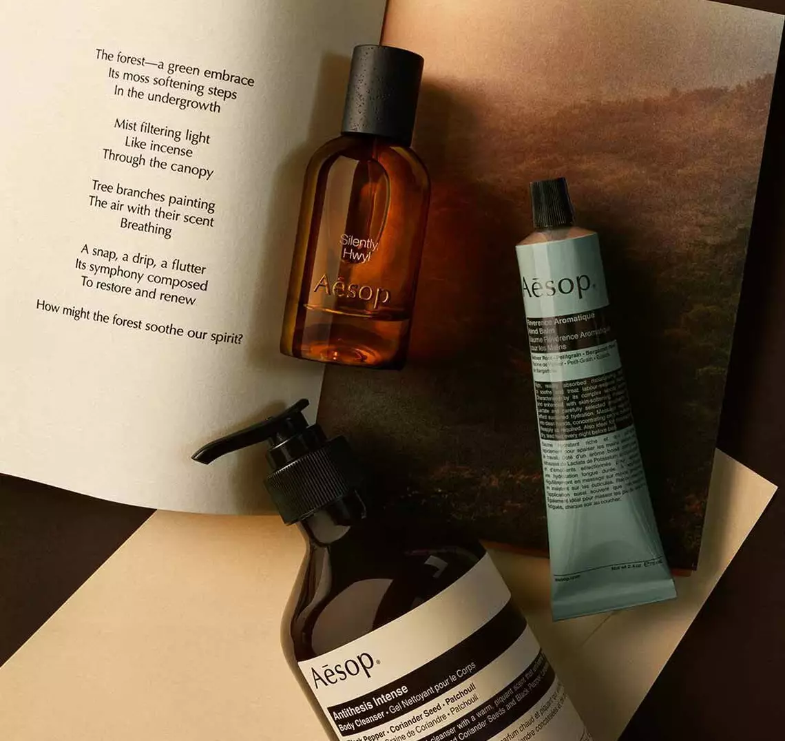 Aesop Silently, Hwyl fragrance bottle, large pump bottle and cosmetic tube on an open book with a woodland photograph.