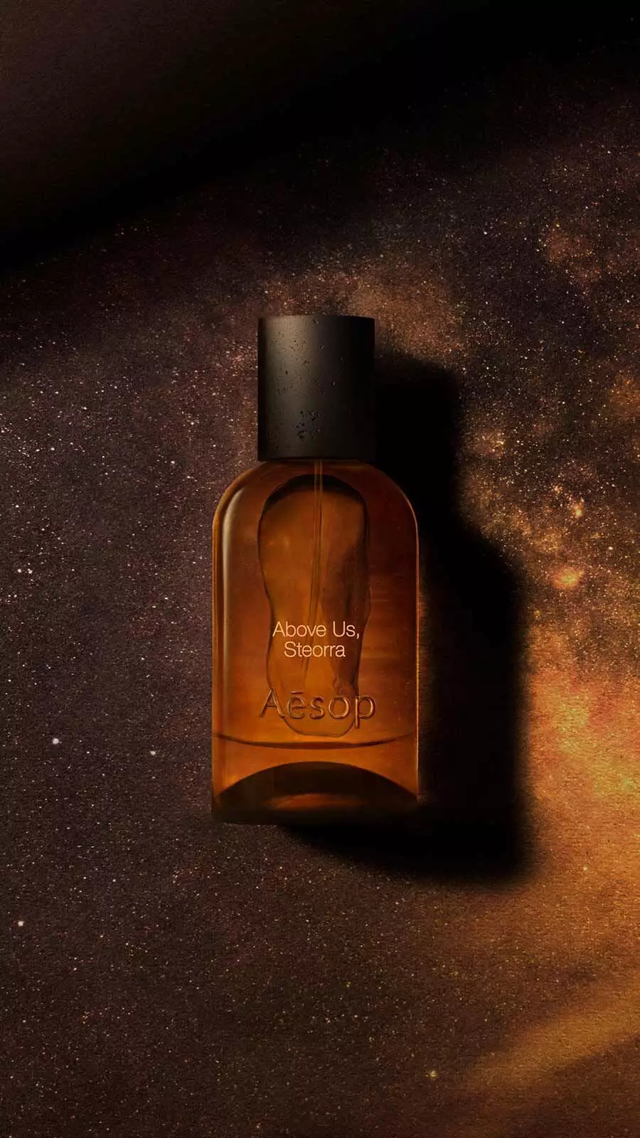 Aesop Above Us, Steorra Eau de Parfum bottle floating against a dark amber background with nebula haze and cosmic light.
