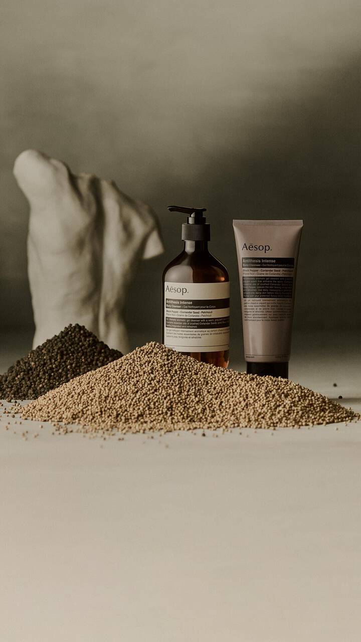 Aesop Antithesis Intense Body Cleanser pump bottle and tube beside piles of black peppercorns and coriander seeds.