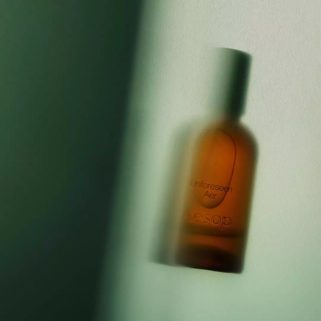 A softly blurred amber Aesop Unforeseen Aer bottle with a dark cap appears against a pale green background and deep green shadow.