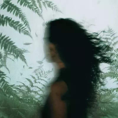 A blurred person in a sleeveless black top, with long dark hair swept sideways, appears against green fern imagery.