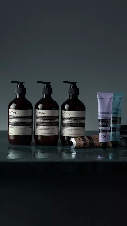 Three Aesop pump bottles, two tubes, and a small amber bottle on a dark reflective surface.