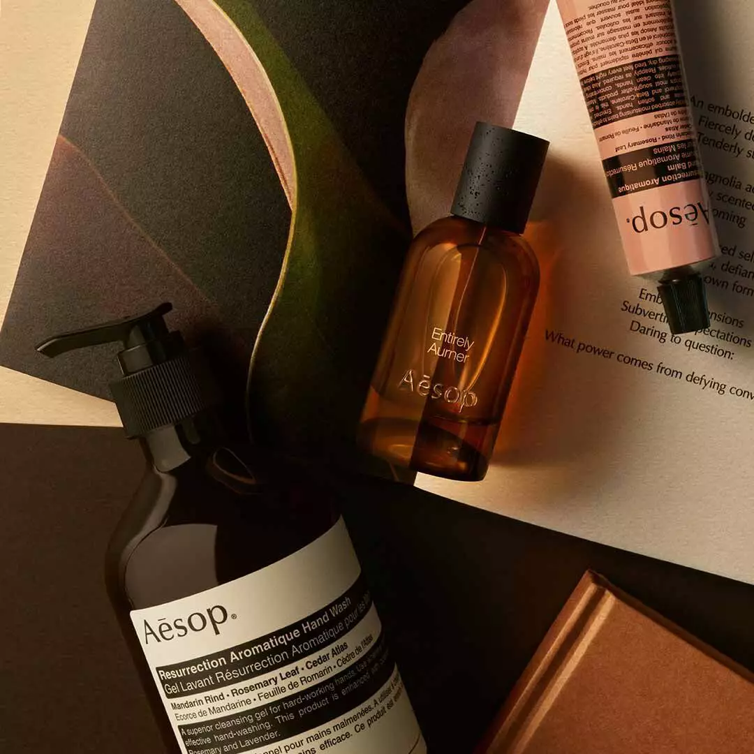 Aesop Entirely Aurner bottle with pump bottle and peach tube arranged on papers and curved dark shapes.