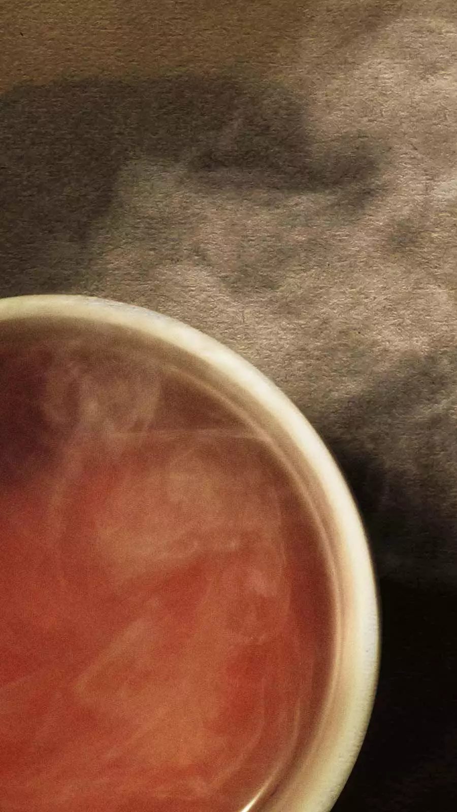 Small ceramic bowl filled with warm red-orange liquid with wisps of steam against a soft grey background.