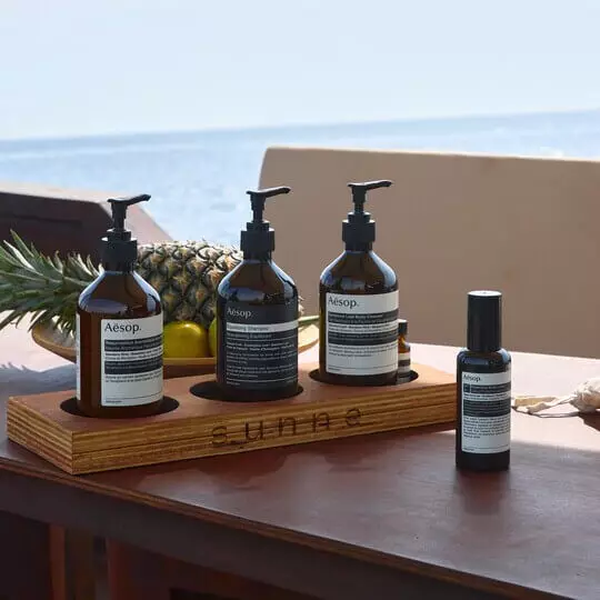 Five Aesop formulations and tropical fruit on a table by the beach.