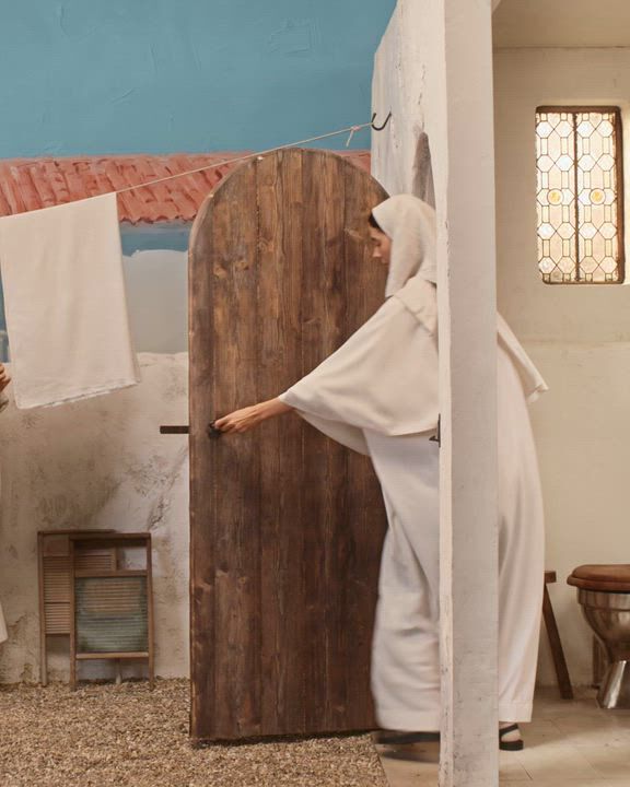 Person in a white robe entering a rustic outdoor-style bathroom with a wooden door and toilet