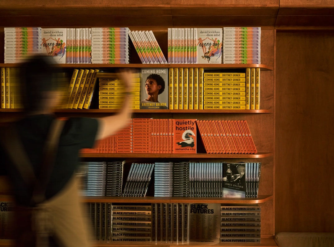 Blurred person walking past warm wooden shelves filled with yellow, orange, and red books.