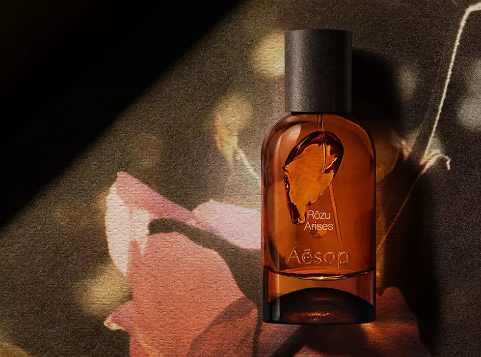 Aesop Rozu Arises amber fragrance bottle on a dark surface beside a pink botanical illustration and pale root.