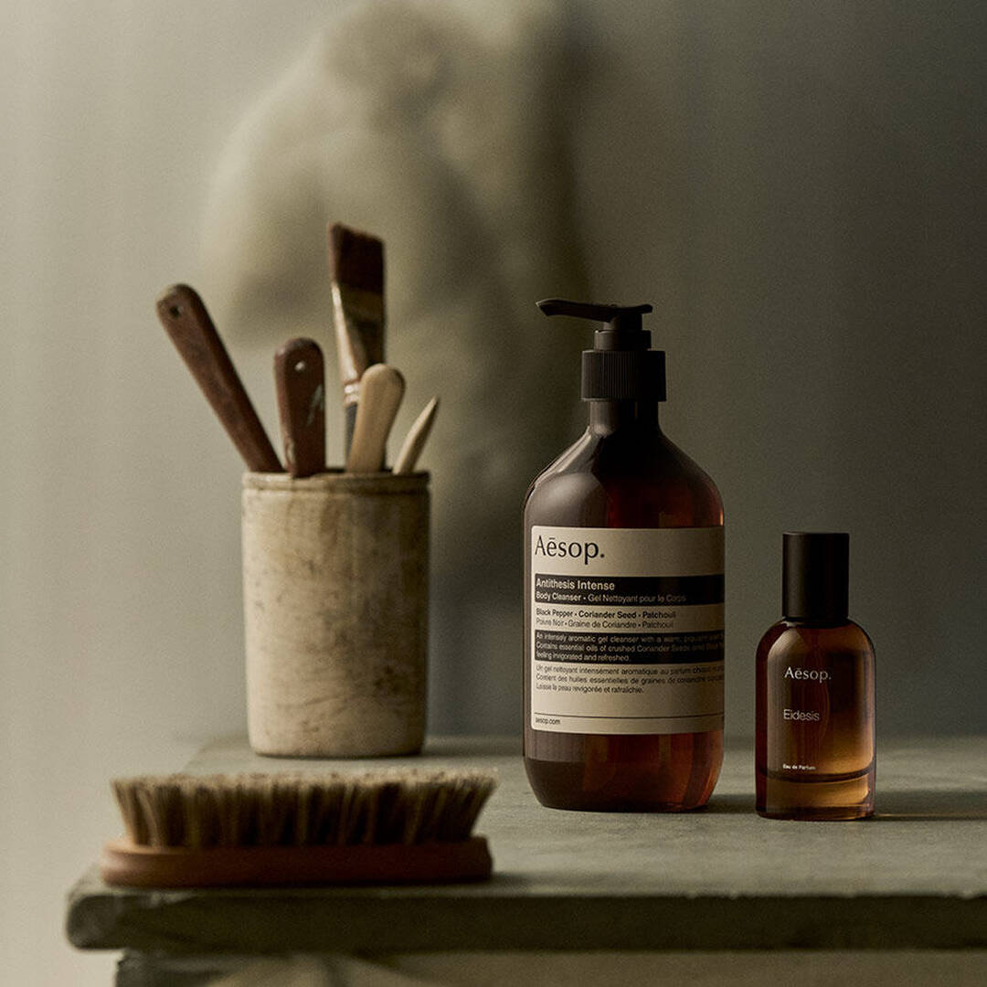 Aesop Antithesis Intense Body Cleanser pump bottle and Eidesis fragrance bottle beside brushes on a shelf.