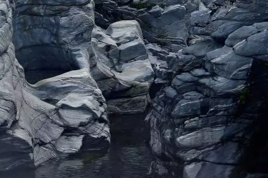 Layered slate-grey rock formations above dark water, showing striated mineral textures, cracks, and naturally sculpted contours.