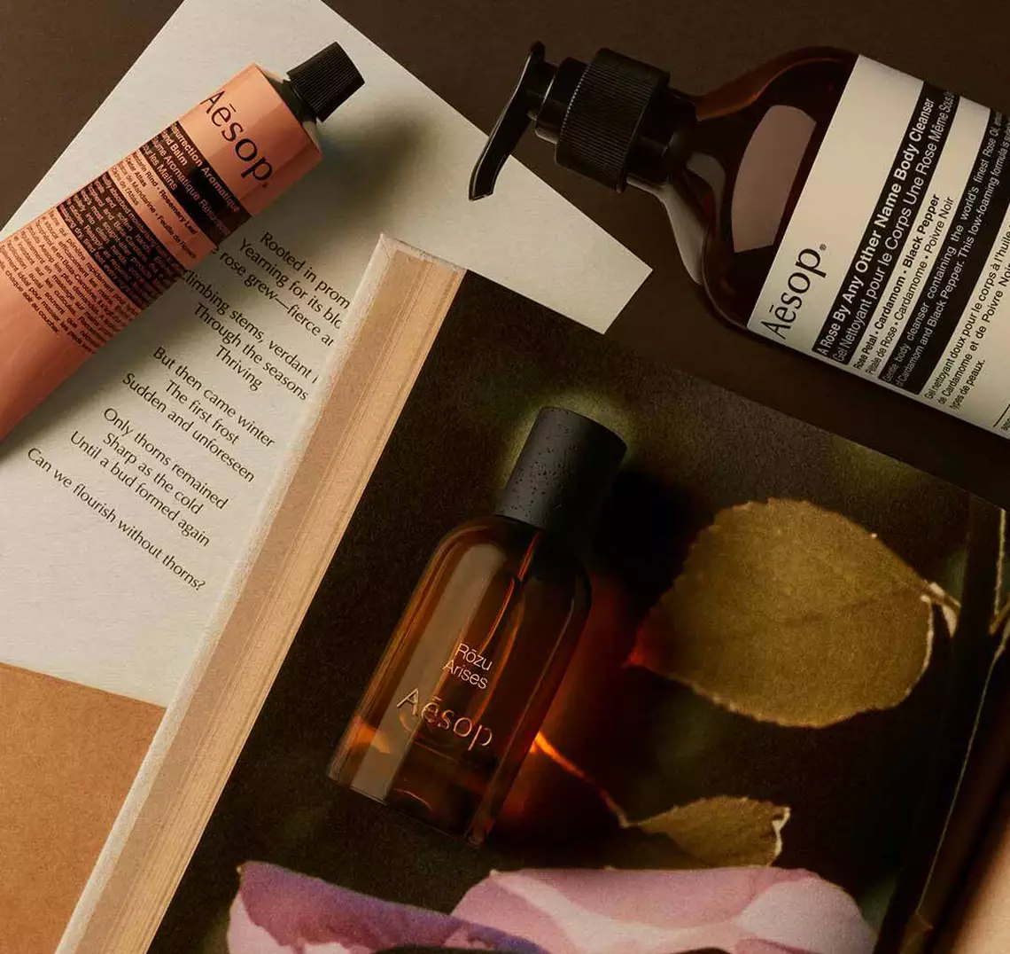 Aesop Rozu Arises fragrance bottle, large pump bottle and mist spray bottle on an open book with a pink rose petal.