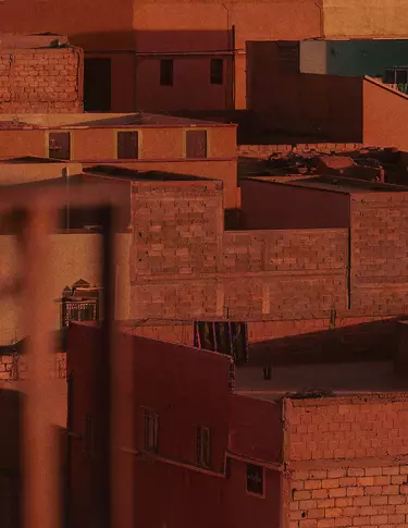 Terracotta city rooftops and walls in warm rust-red light, with blocky architecture, shadowed facades, and textured masonry surfaces.