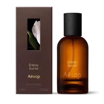 Amber glass Aesop Entirely Aurner Eau de Parfum bottle beside its brown outer box on a white background.