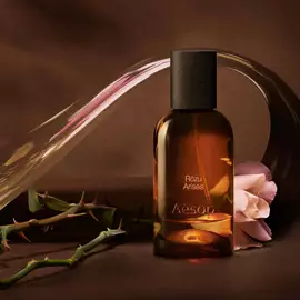 Aesop Rozu Arises bottle with a rose stem and flower petals on a warm surface.