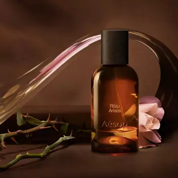 Aesop Rozu Arises bottle with a rose stem and flower petals on a warm surface.