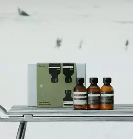 Three amber Aesop bottles with black caps stand beside a green and silver box on a metal sledge in snow.