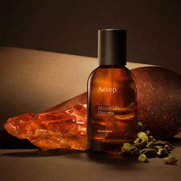 Aesop Above Us, Steorra bottle beside an amber resin piece and green cardamom pods on a warm brown surface.