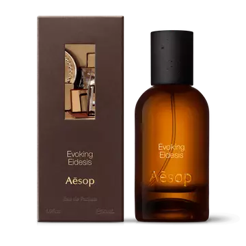 Amber glass Aesop Evoking Eidesis Eau de Parfum bottle beside its brown outer box on a white background.